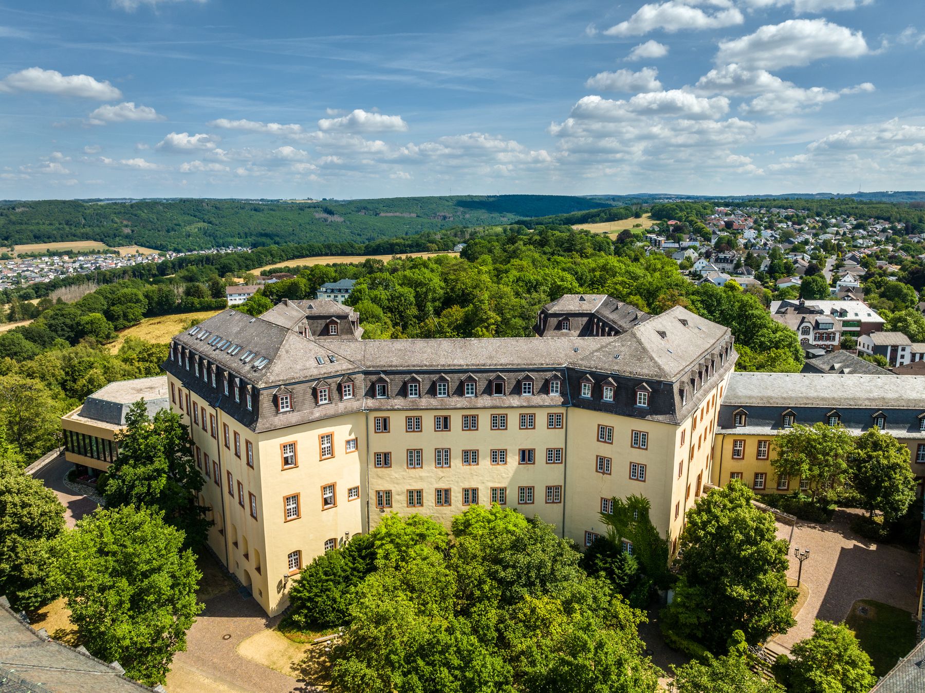 Ein großes, historisches, gelbes Gebäude mit vielen Fenstern und einem grauen Dach, umgeben von üppigen grünen Bäumen, steht auf einem Hügel mit Blick auf eine kleine Stadt und eine hügelige Landschaft unter einem blauen Himmel mit vereinzelten Wolken.